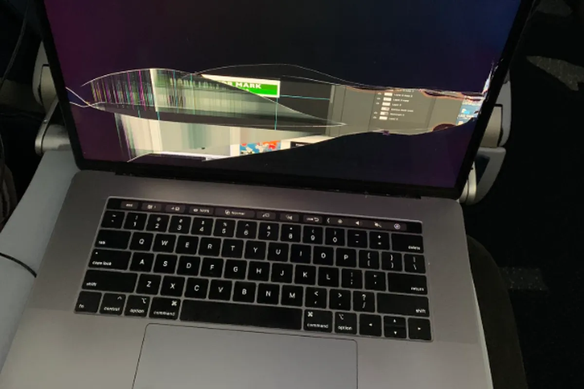 A MacBook Pro with a shattered display, the cracked panel still lit and showing torn image across the break